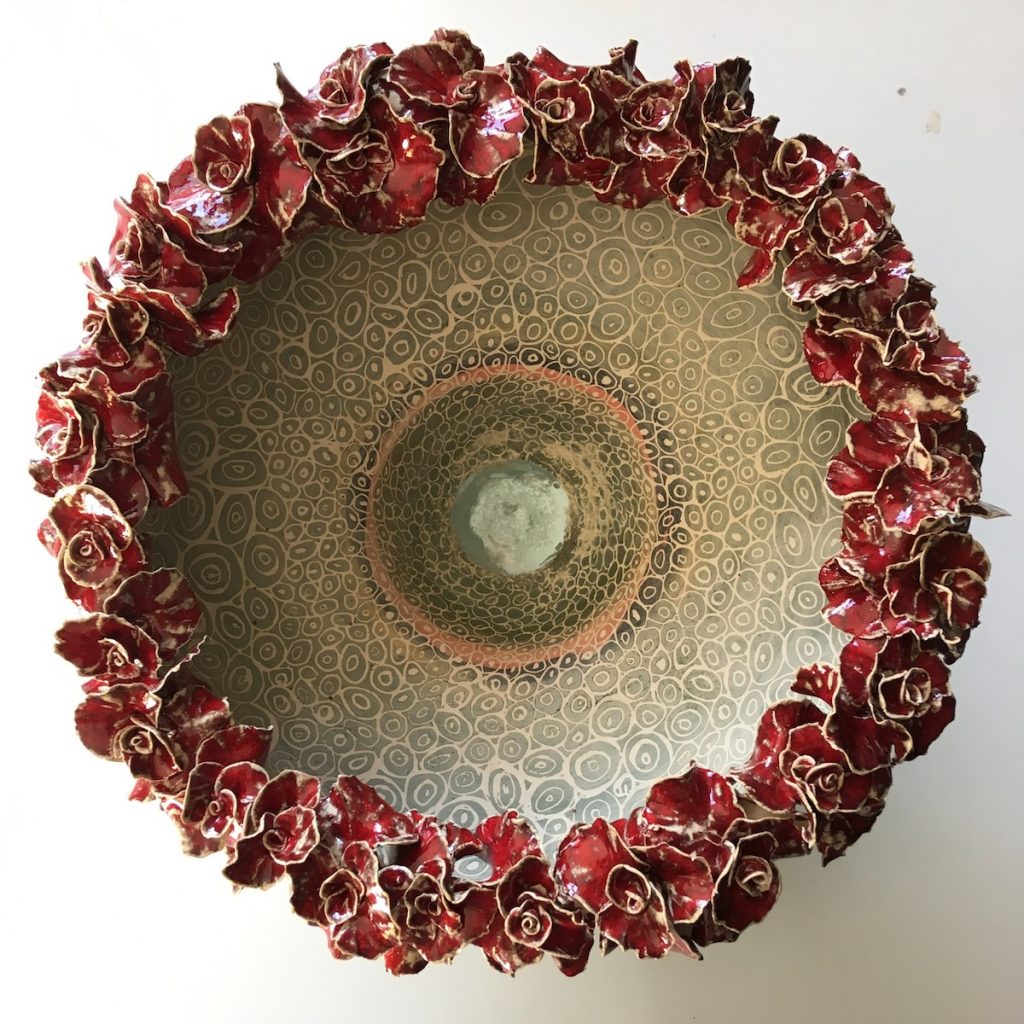 Keramiek schaalvorm. Aardewerk opgebouwd uit ringen met een rand van handgevormde bloemvormen. Bewerkt in meerdere lagen met engobes en glazuren.