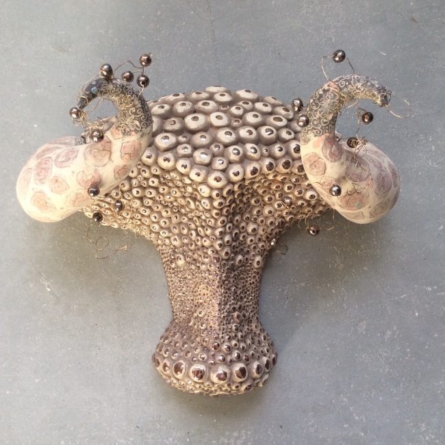Keramiek kunst voor aan de wand. Aardewerk opgebouwd uit ringen en bezet met handgedraaide bolletjes. Bewerkt met engobes, glazuur en glazuurpotlood. Aardewerk kralen aan koperdraad